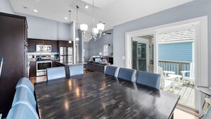 A modern kitchen with dark cabinetry, a central island, and a dining area with a dark wooden table and blue chairs. - Pier Pressure At SandCastle Nags Head Village, Private Pool, On the Golf Course (Nags Head)