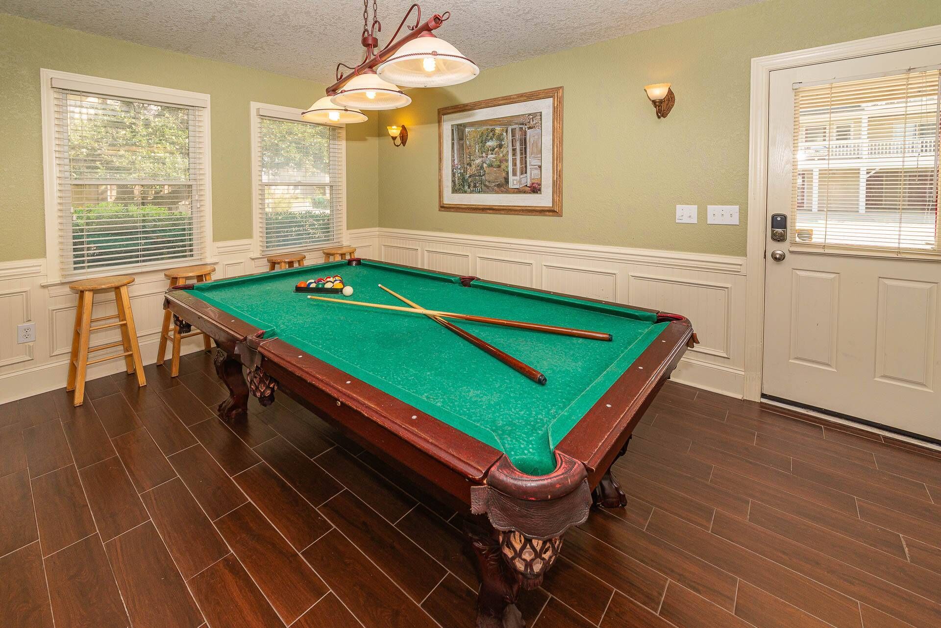 Une table de billard avec des queues et des billes, des tabourets en bois, un tableau encadré et une porte avec une fenêtre.