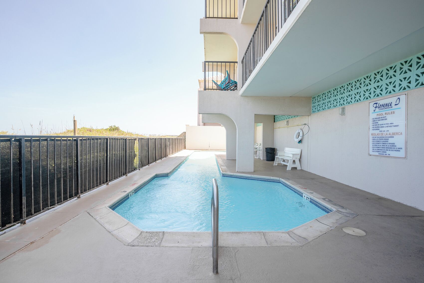 Una piscina con barandilla metálica, un banco blanco y un cartel con normas de la piscina.