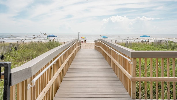 Pasarela de madera que conduce a una playa con sombrillas y una valla.