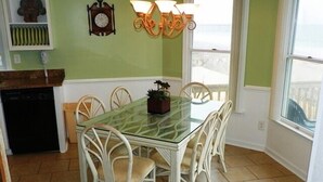 A dining area with a glass-top table surrounded by white chairs, a clock on the wall, and a window with a view. - KH5203- Oceanfront With Two Masters, Hot Tub, Covered Porch and Dog Friendly! (Kitty Hawk)