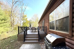 Una terraza de madera con una parrilla y mobiliario exterior, rodeada de árboles.