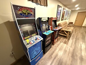 Espaço de jogos com máquinas de arcade, incluindo Street Fighter II Edition, NBA Jam e Caliber. Há um sofá e uma porta ao fundo.