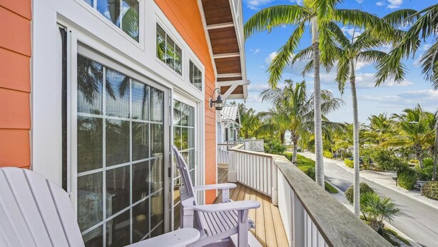 A wooden deck with white Adirondack chairs, a view of palm trees, and a house with orange siding and white trim. - Beautiful Home Two Houses from the Beach!  Pool, Private Patio, 3 Bedrooms (Holmes Beach)
