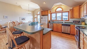 A kitchen with wooden cabinets, a central island, and a dining area with chairs. - Paws and Reflect - Corolla, Ocean Sands, 200 Yds to Beach, Pool, Hot Tub, Pool Table, Pets OK (Corolla)