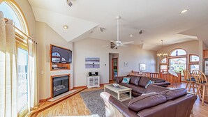A spacious living room with a fireplace, leather sofa, and a dining area with wooden chairs. - Paws and Reflect - Corolla, Ocean Sands, 200 Yds to Beach, Pool, Hot Tub, Pool Table, Pets OK (Corolla)