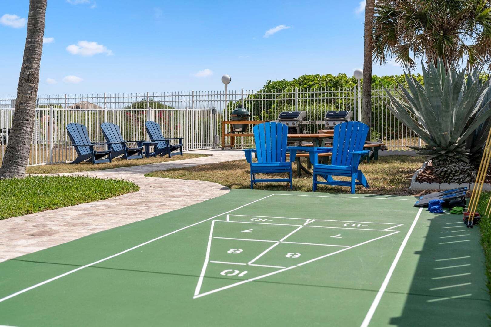 Shuffleboard-pelialue, jossa on siniset tuolit ja pöytä, ja jonka ympärillä on aita ja vihreää kasvillisuutta.