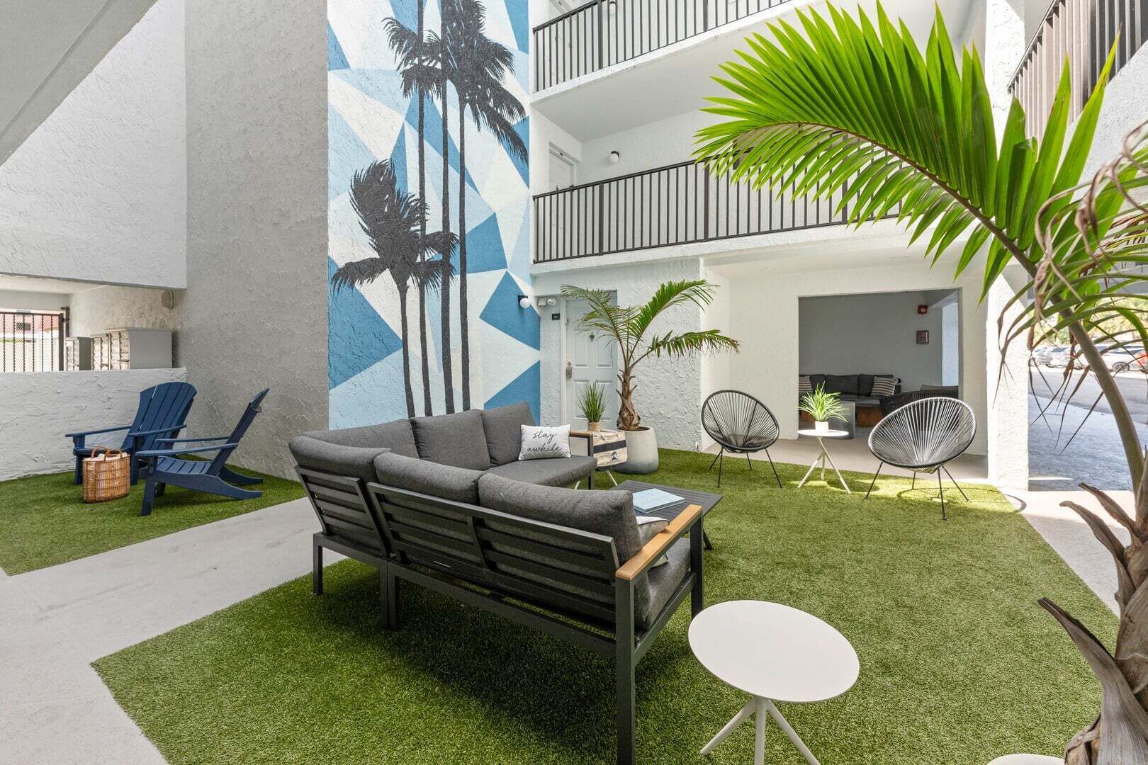 A modern outdoor seating area with a sofa, chairs, and a small table, surrounded by artificial grass and palm plants.