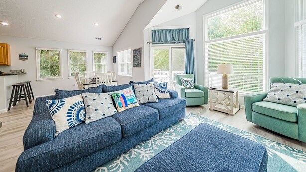 Een ruime woonkamer met een hoge plafon, een blauwe bank, een groene fauteuil en een eethoek met een witte tafel en stoelen.