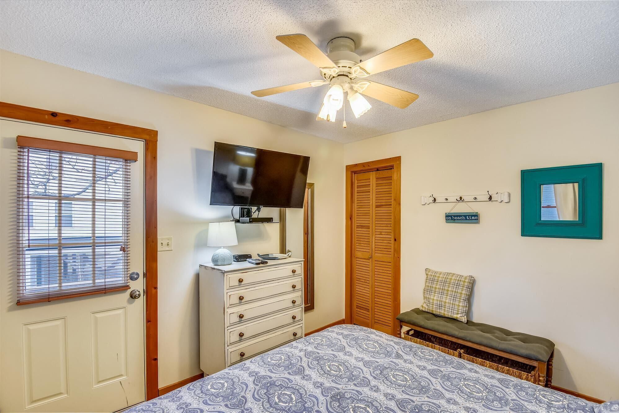 A bedroom with a bed, dresser, television, ceiling fan, and a bench.