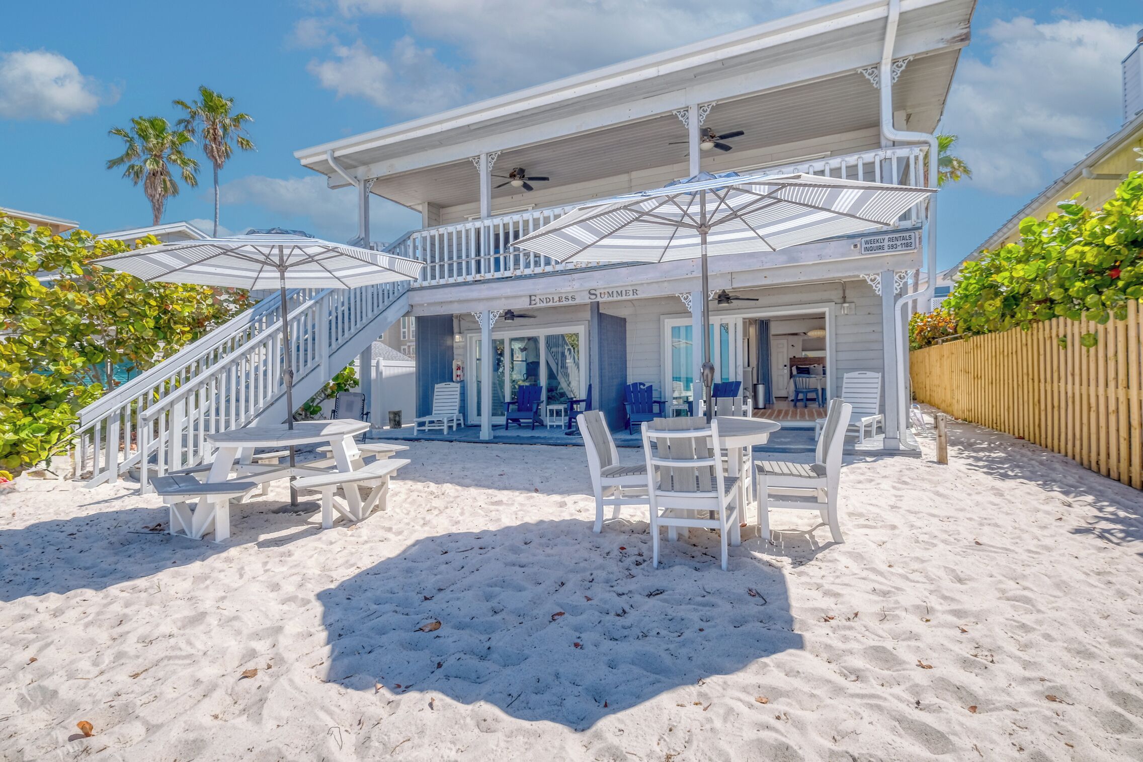 Un bâtiment situé en bord de mer, doté d’un patio couvert, de mobilier d’extérieur blanc et d’un escalier menant à l’entrée.