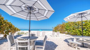 Ett sittområde vid stranden med vita bord och stolar, randiga parasoller och utsikt över havet.