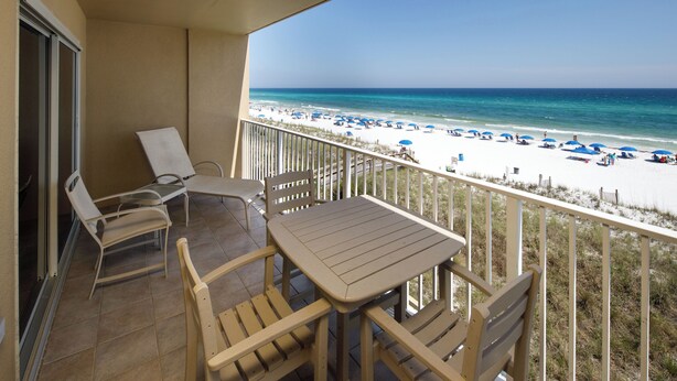Een balkon met een tafel en stoelen, uitkijkend op een strand met parasols en mensen.