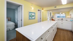 Een lichte keuken met witte kastjes en een houten vloer, een deur naar een slaapkamer, en een raam met uitzicht.