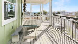 A covered outdoor deck with a wooden table and bench, white railings, and a view of the ocean and other buildings. - Directly Next to Community Pool! Beach view! ~Fried Green Tomatoes (Cape San Blas)