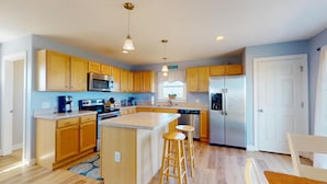 A modern kitchen with wooden cabinets, stainless steel appliances, and a central island with stools. - Summer Paws | 3BR 4x4 OBX | Hot Tub | Pets | 4 night stay available for fall (Carova)