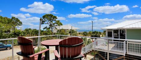 A deck with two Adirondack chairs and a small table, overlooking a body of water and a building with a white roof.