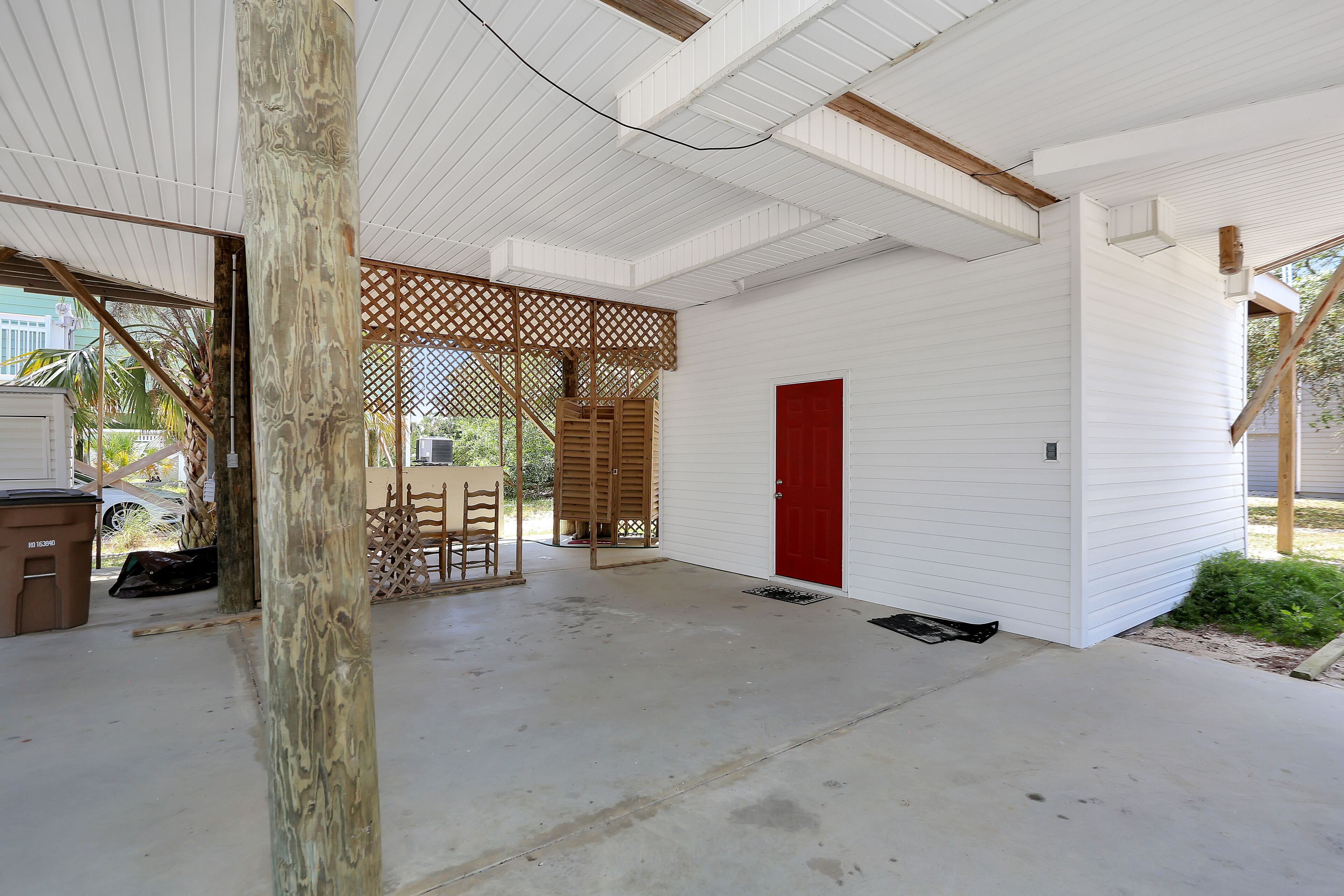 A covered outdoor area with a concrete floor, a wooden table and chairs, a red door, and a lattice fence.