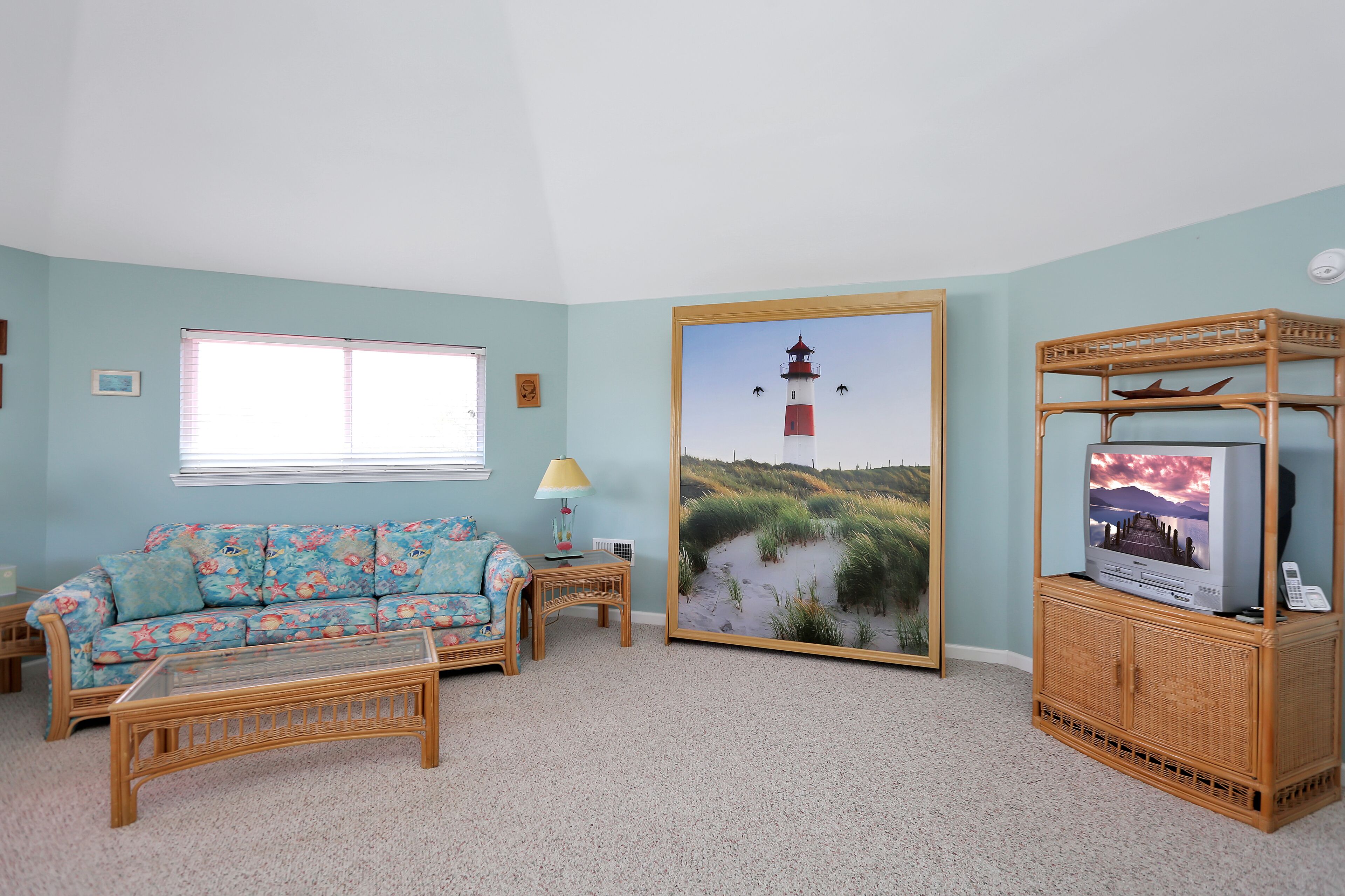 Una sala de estar con un sofá, una mesa de centro, un televisor sobre un mueble bajo y un cuadro con una imagen de un faro.