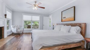 A bedroom with a large bed, a ceiling fan, a fireplace, a window with a view, and a painting on the wall. - On the Creek, Sleeps 10, Close to Town - Perfect Wimberley Escape (Wimberley)