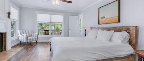 A bedroom with a large bed, a ceiling fan, a fireplace, a window with a view, and a painting on the wall.