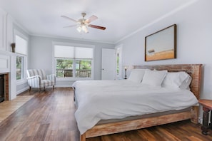 A bedroom with a large bed, a ceiling fan, a fireplace, a window with a view, and a painting on the wall.