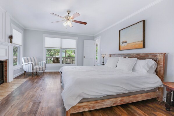 A bedroom with a large bed, a ceiling fan, a fireplace, a window with a view, and a painting on the wall.