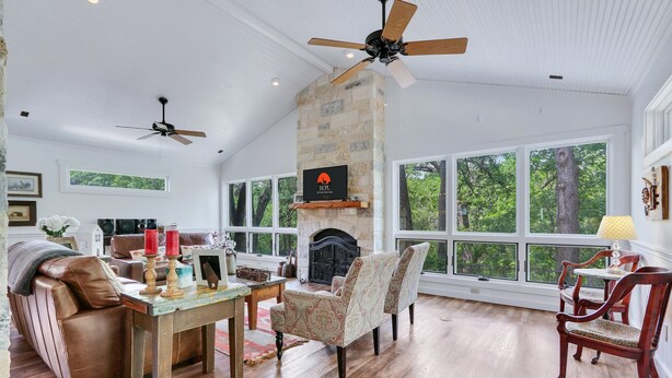 A spacious living room with a fireplace, large windows, and a ceiling fan. - On the Creek, Sleeps 10, Close to Town - Perfect Wimberley Escape (Wimberley)
