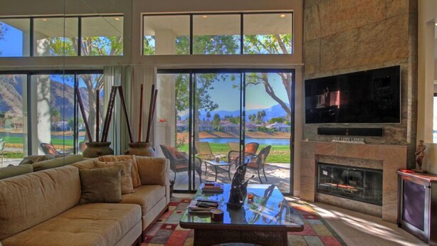 A spacious living room with a large sofa, a coffee table, and a fireplace. There are large windows offering a view of the outdoors. - Delightful 3 Bed Home with Amazing Views LQ171 LIC#110324 (La Quinta)