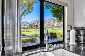 A kitchen with a sink, a window with a view of a golf course and mountains, and a coffee maker.