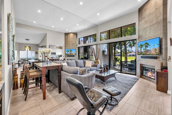 A spacious living area with a fireplace, large windows, and a kitchen in the background.