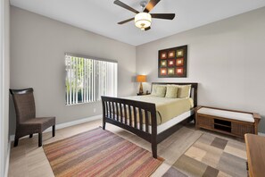 Un dormitorio con cama, mesita de noche, silla, ventana con persianas, ventilador de techo y una obra de arte en la pared.