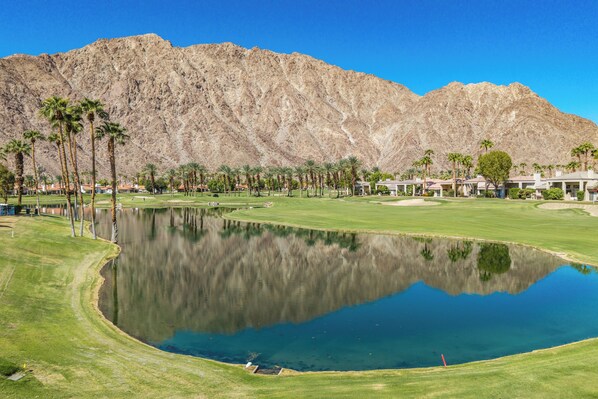 Campo de golf con un estanque, palmeras y montañas al fondo.