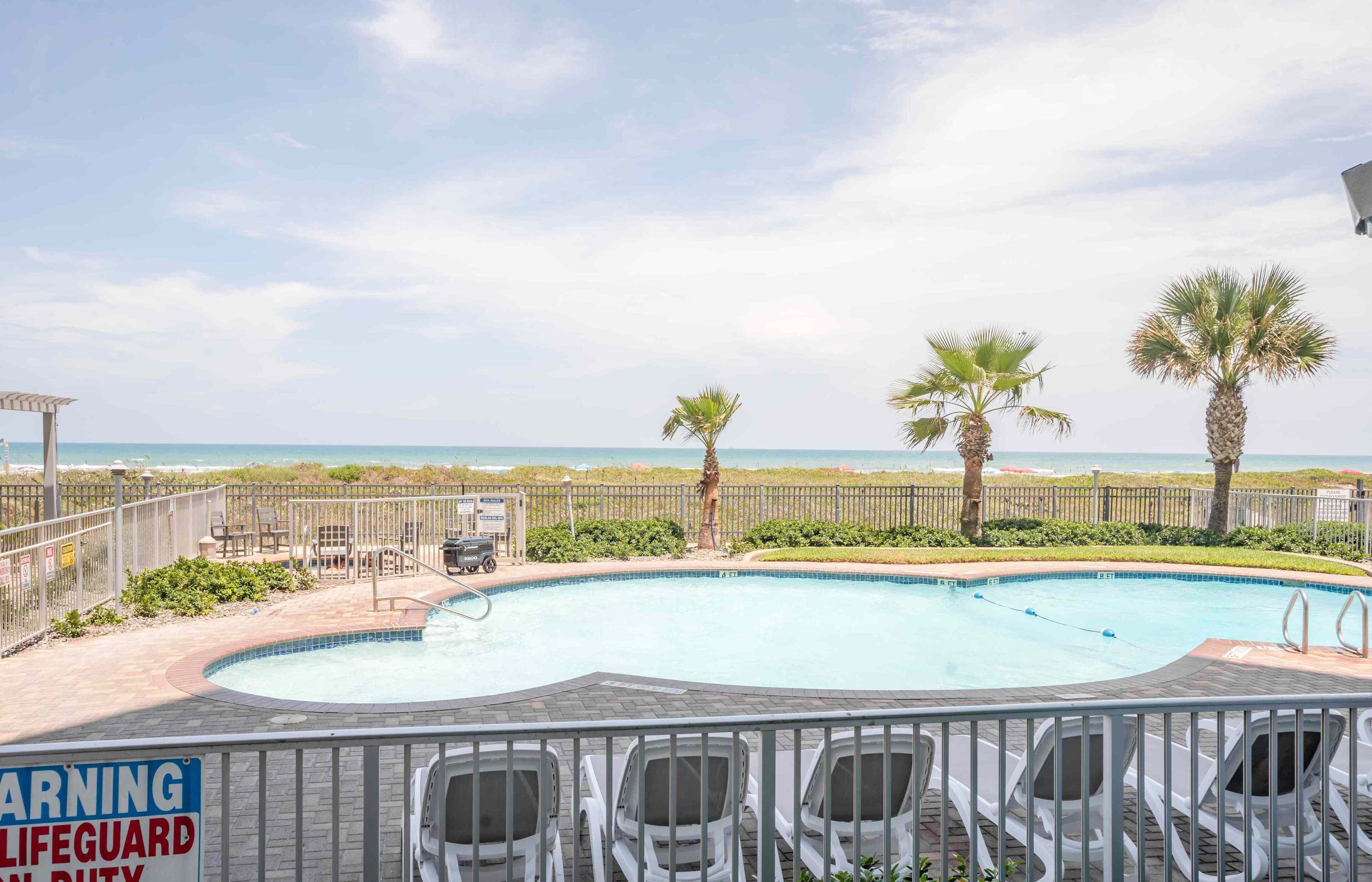 A swimming pool with a lifeguard warning sign, a beach view, and a fenced area with chairs.