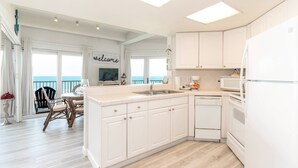 Una cocina moderna con armarios blancos, horno empotrado y lavavajillas. Cuenta con una zona de comedor con mesa y sillas, y vistas al océano a través de amplios ventanales.