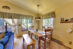 A cozy dining area with a wooden table, chairs, and a bench. There is a blue couch, a window with curtains, and a wall-mounted shelf with a bear decoration.