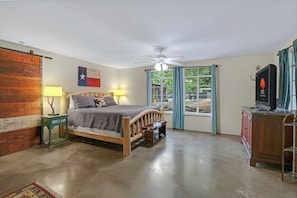 Un dormitorio con cama, mesita de noche, lámpara, ventilador de techo, ventana con cortinas y televisor sobre un mueble.
