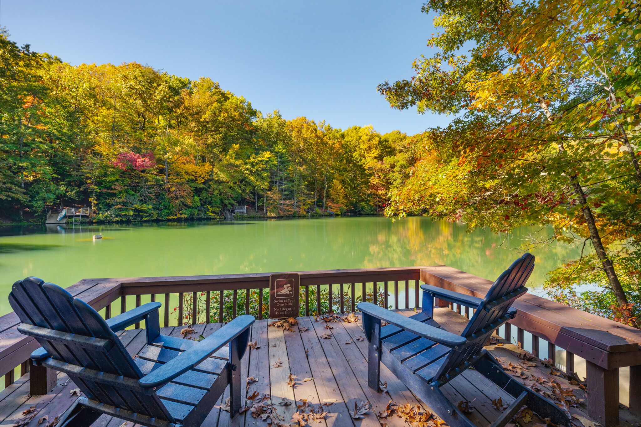 Eine Holzterrasse mit Adirondack-Stühlen mit Blick auf einen ruhigen See, umgeben von Herbstlaub.