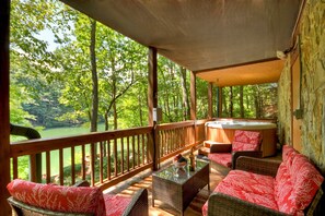 A covered outdoor area with wicker furniture, a hot tub, and a view of a forested area. - Embrace serenity at Morningstar on the Lake, this pet-friendly haven features a soothing hot tub, custom firepit, fishing dock, canoe, hiking trails, and WiFi (Cherry Log)