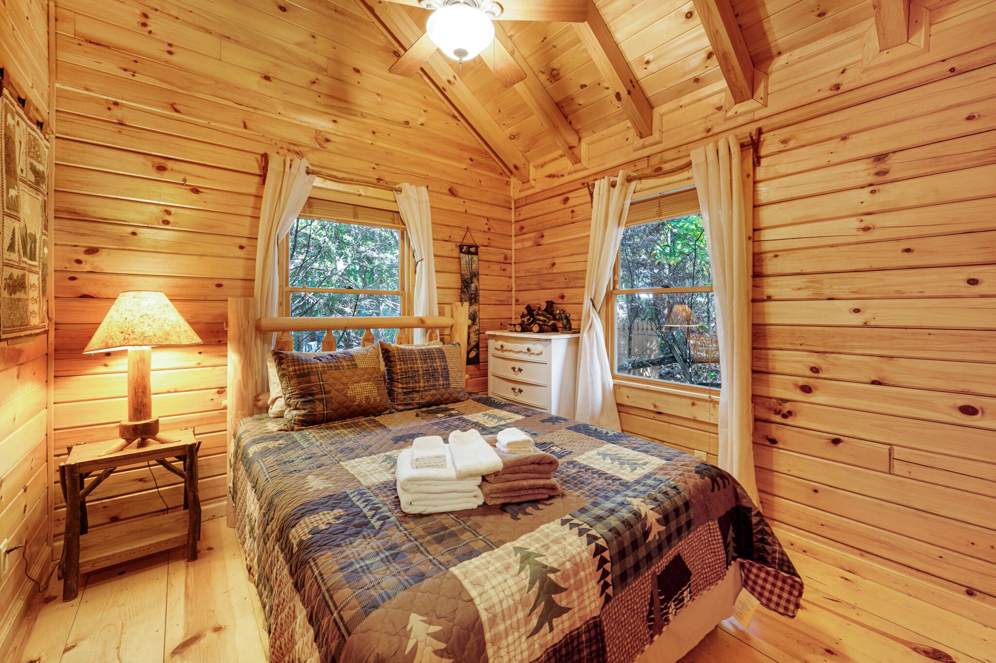 Un dormitori de cabana de fusta amb llit, lampadeta de nit, tocador i finestres.