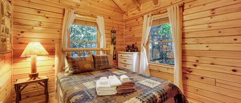 Un dormitori de cabana de fusta amb llit, lampadeta de nit, tocador i finestres.