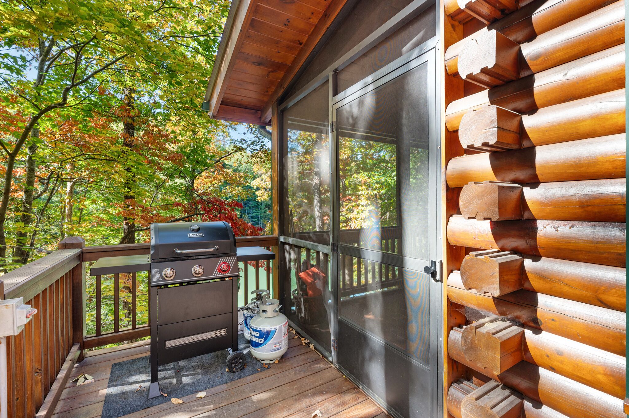 A wooden deck with a grill and propane tank, surrounded by autumn foliage.