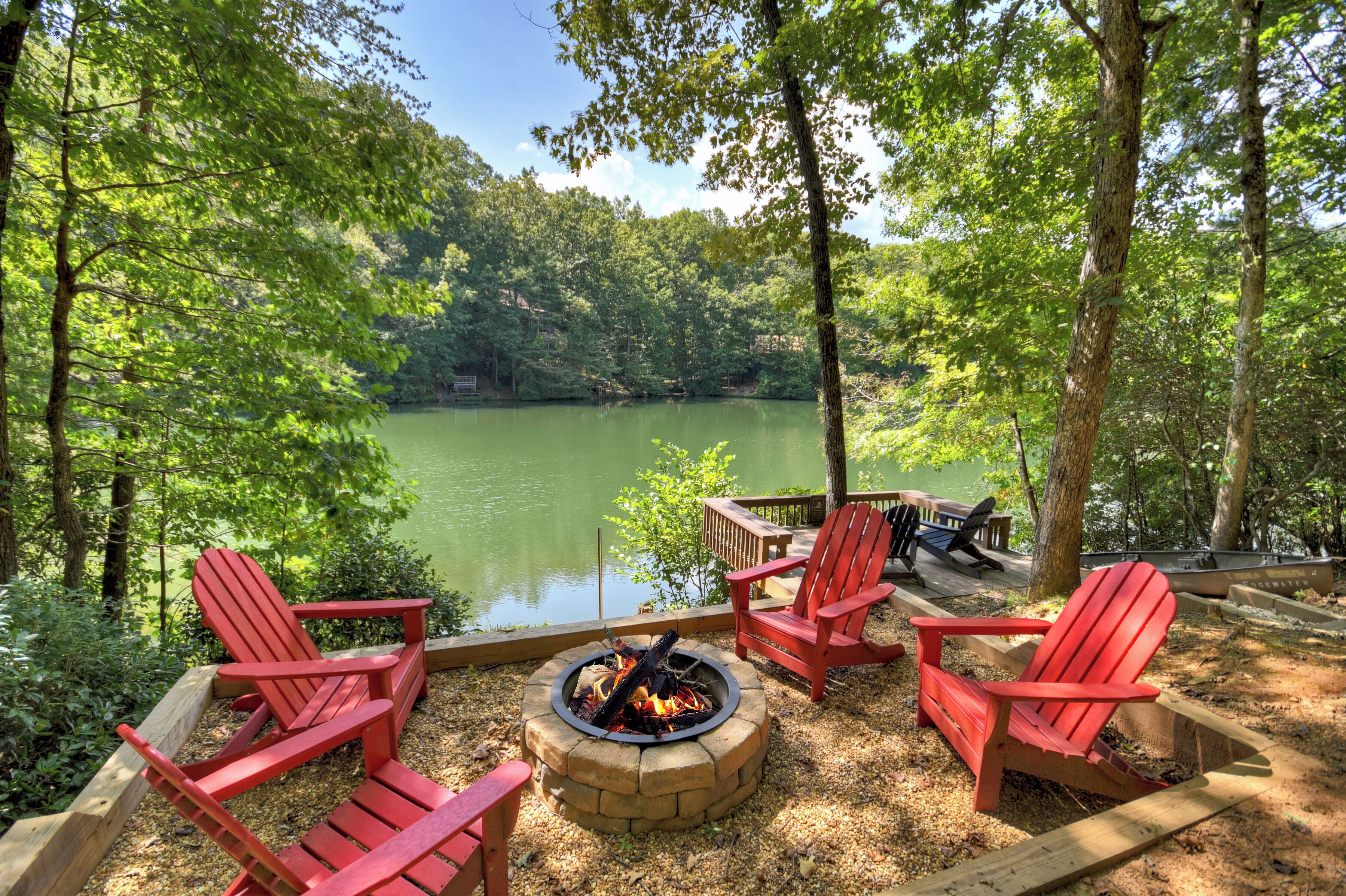 A lakeside deck with red Adirondack chairs, a fire pit, and a wooden deck overlooking a lake.