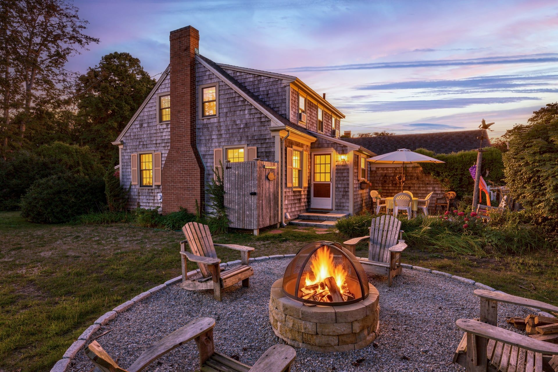 A two-story house with a fire pit in the front yard, surrounded by wooden chairs.