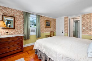 Een slaapkamer met een bed, een dressoir en een stoel. Er is een raam met gordijnen, een deur en een schilderij aan de muur.