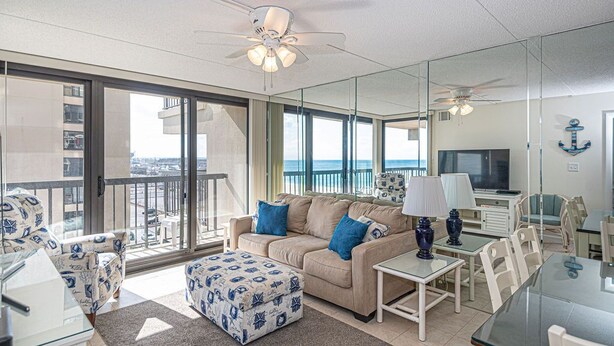 A modern living room with a sofa, ottoman, and a dining area with a TV and lamp. - Comfortable, cozy 3-bedroom oceanfront condo with free WiFi, a breathtaking ocean view, an indoor and outdoor pool, and more located uptown and mere steps to the beach! (Ocean City)