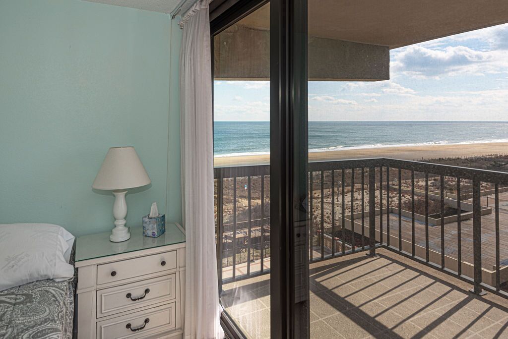 A room with a view of the beach, a bedside table with a lamp, and a window with a view of the ocean.