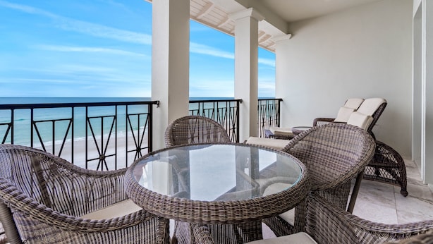 A balcony with wicker furniture and a glass table overlooking the ocean.