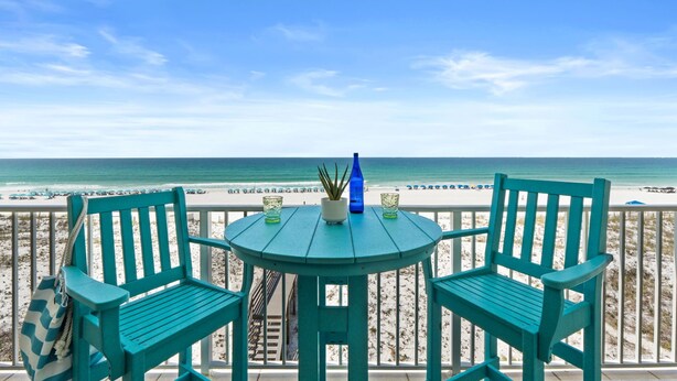 Un balcon avec deux chaises vertes et une petite table, donnant sur une plage sous un ciel dégagé.