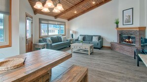 A spacious living room with a high ceiling, a fireplace, a large window, and a wooden dining table.